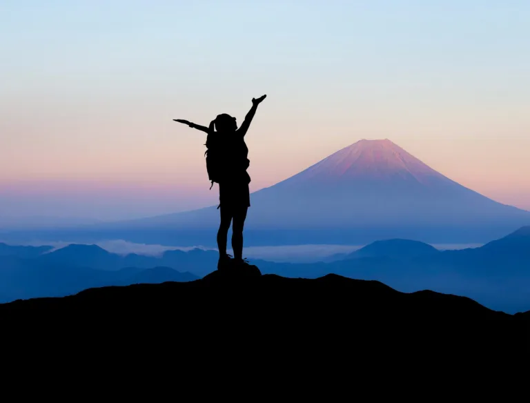Silhouette of a hiker with arms raised in victory against the backdrop of a mountain at sunrise, embodying the spirit to challenging yourself.