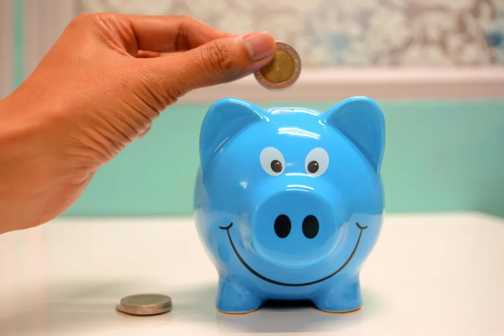 A hand placing a coin into a smiling blue piggy bank, representing the first step in investing and building wealth