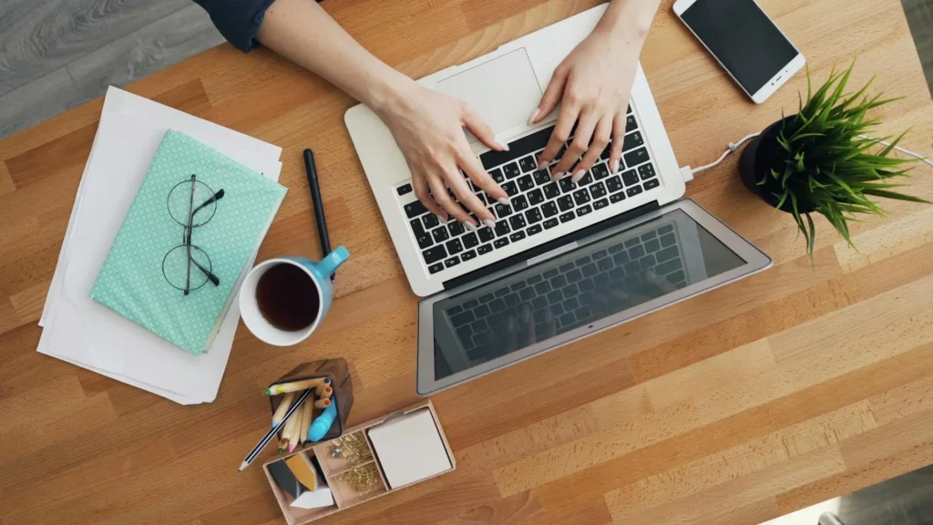A person working on a laptop at a neatly organized desk with a notebook, glasses, and a cup of tea.