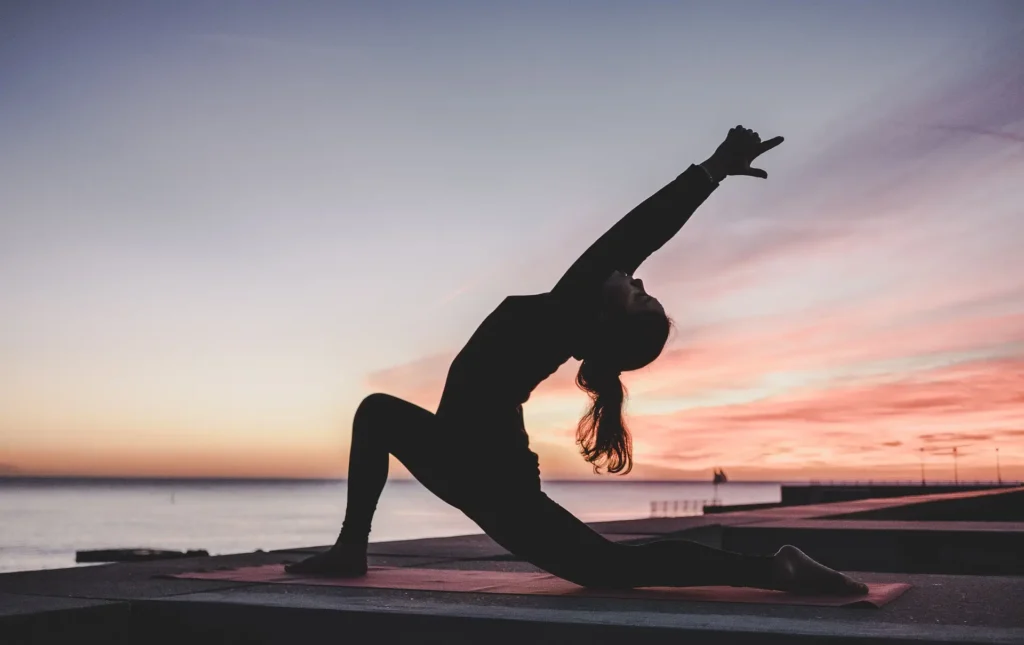 Person practicing yoga at sunset near the water, exemplifying fitness while traveling.