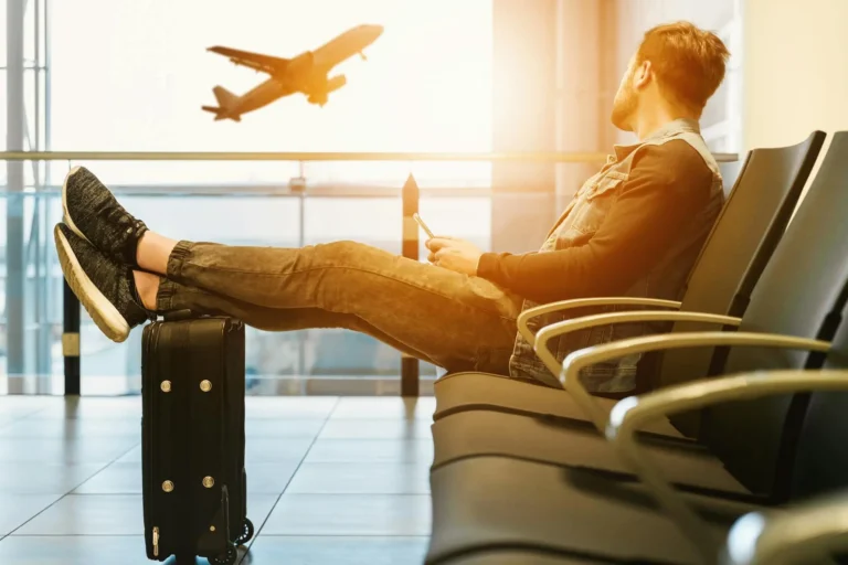 Traveler sitting in an airport lounge with feet on suitcase, watching a plane take off during sunset with prepared all item from summer travel checklist.