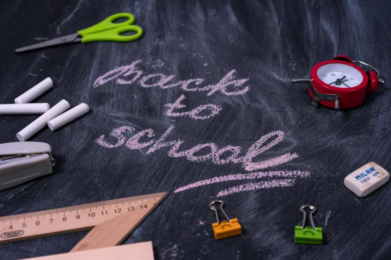 Back to school essentials on a chalkboard with supplies including chalk, a ruler, scissors, and an alarm clock.